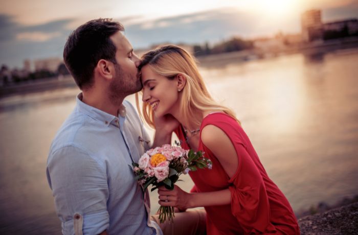 Foto de una pareja frente a un lago. El hombre besa en la frente a la mujer, mientras ella sostiene unas flores.