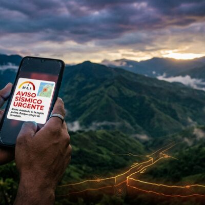 Manos sosteniendo un celular con una alerta de sismo urgente de magnitud 6.1, con montañas de fondo