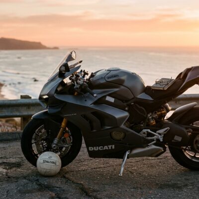 Motocicleta Ducati gris estacionada en una carretera costera al atardecer con un balón de voleibol junto a la rueda